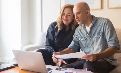 A smiling couple holding papers and going over their business’s finances on a laptop.